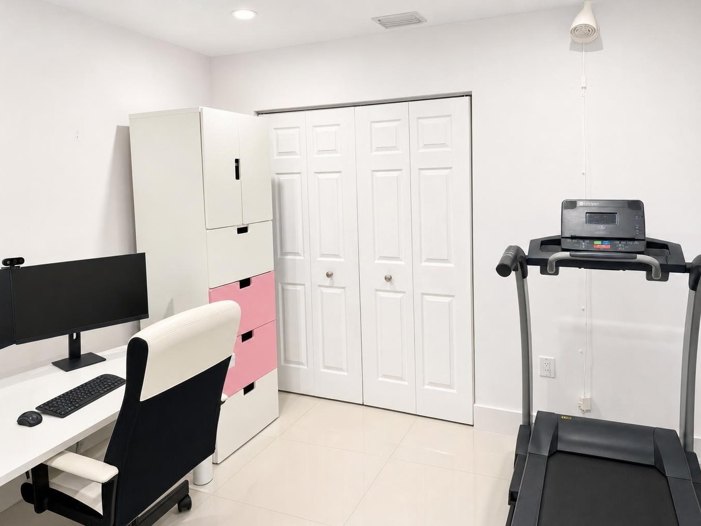 A modern office with a black and white chair, white desk with monitor, pink and white cabinet, and a treadmill against a white wall.