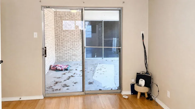 Empty living room with light wood floor and glass sliding door overlooking small brick patio with leaves scattered outside.