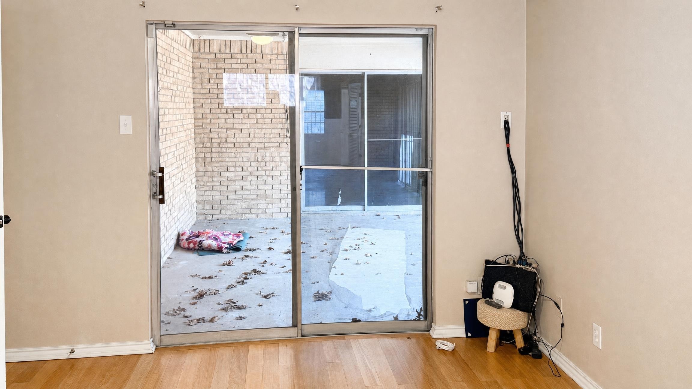 Empty living room with light wood floor and glass sliding door overlooking small brick patio with leaves scattered outside.