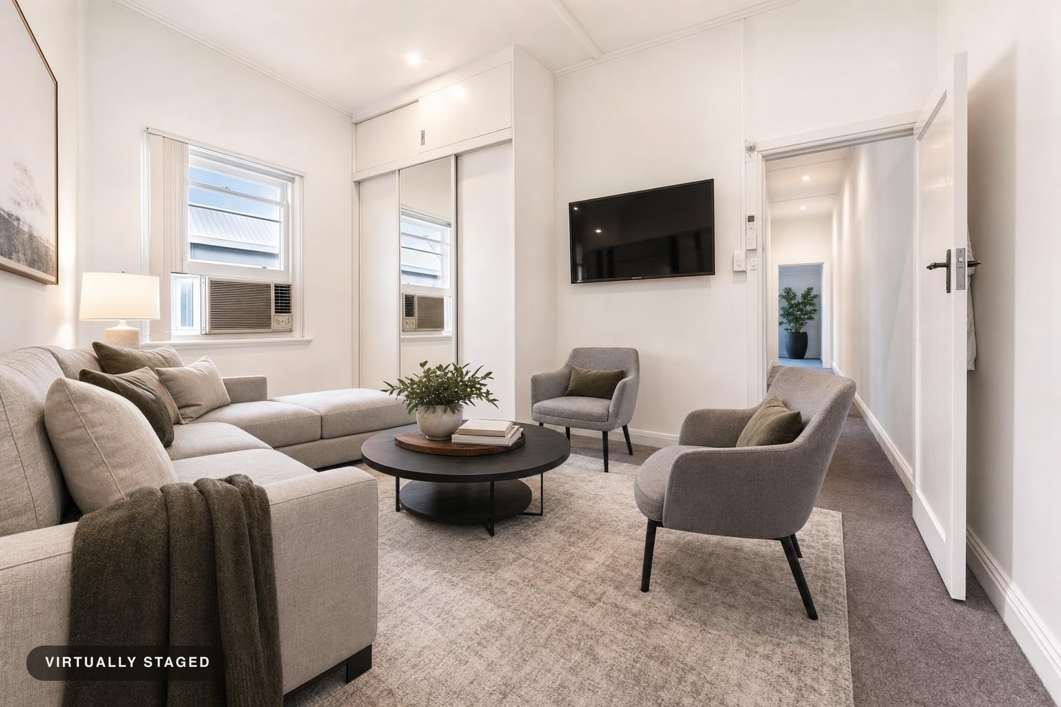 Modern living room with light gray sectional sofa, two matching armchairs, a round black coffee table, and a flat screen TV on a white wall.