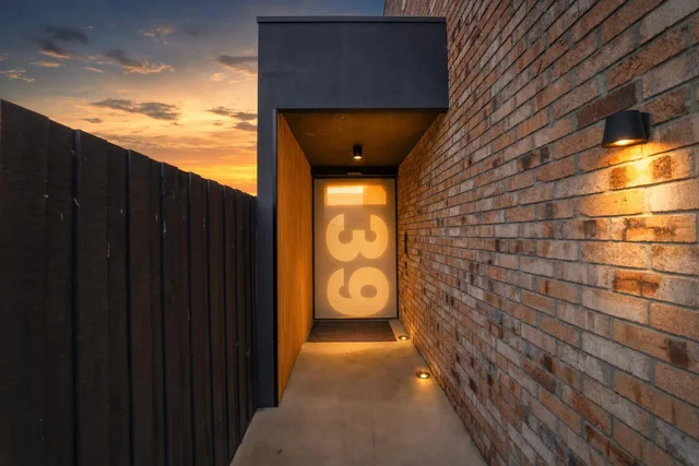 Narrow exterior hallway at dusk with a brick wall on the right, wooden fence on the left, and a door with large illuminated numbers 139.