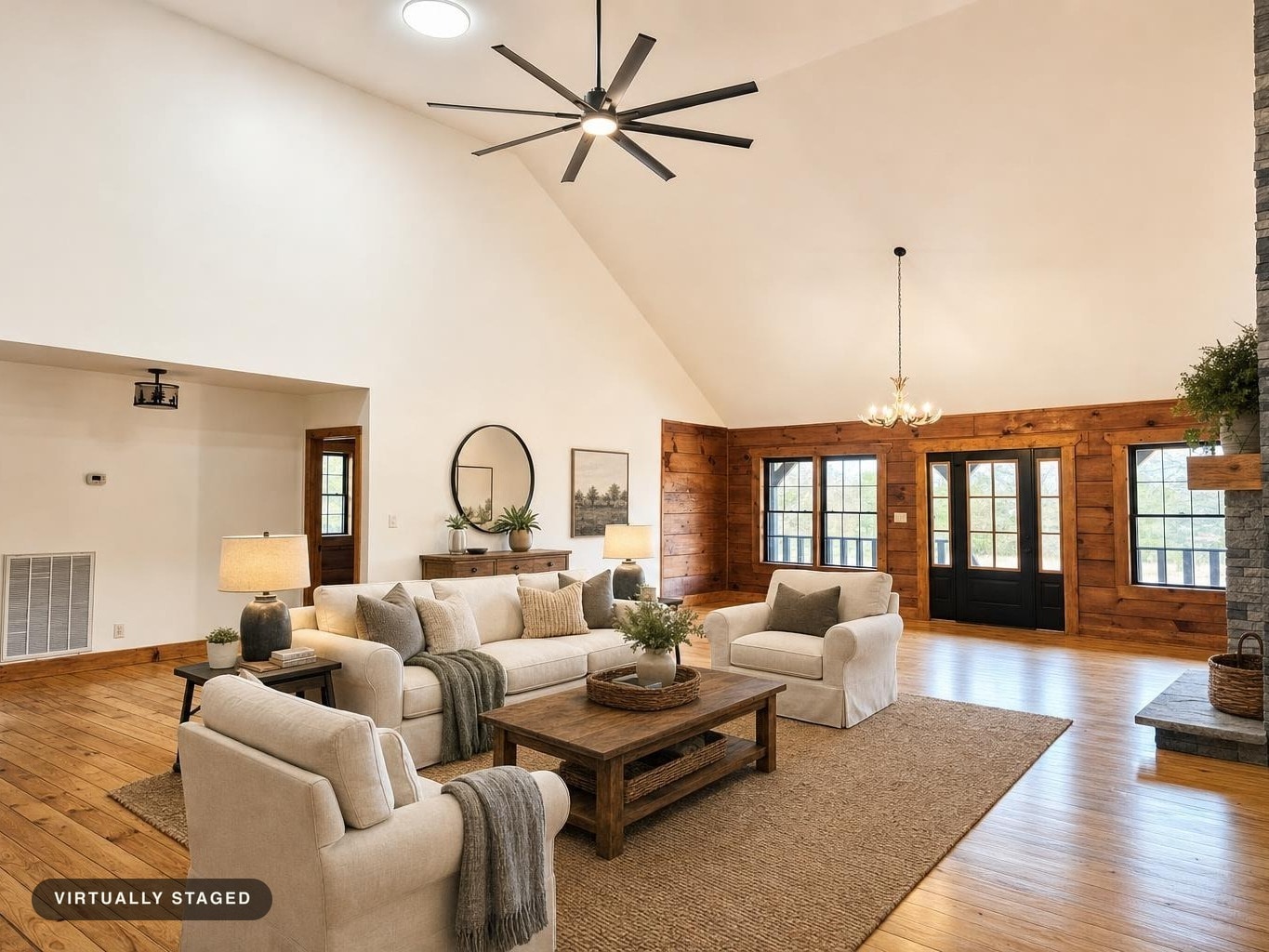 Spacious living room with vaulted ceiling, beige sofa and chairs, wooden coffee table, and wood-paneled walls with large windows.