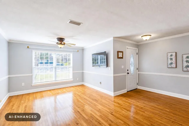Empty living room with gray walls, large window, ceiling fan, wooden floor, front door, and TV mounted on wall.