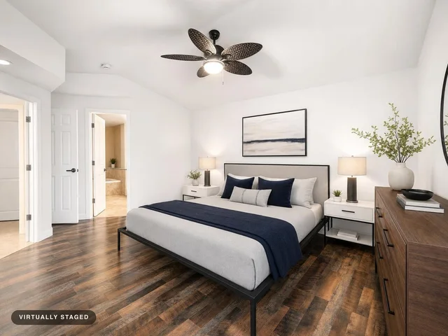 Modern bedroom with a gray upholstered bed, navy blue accents, white nightstands, and wooden dresser on dark wood flooring.