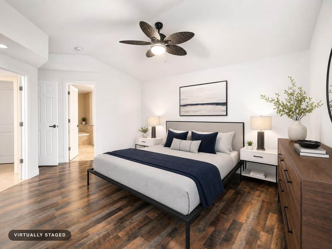 Modern bedroom with a gray upholstered bed, navy blue accents, white nightstands, and wooden dresser on dark wood flooring.