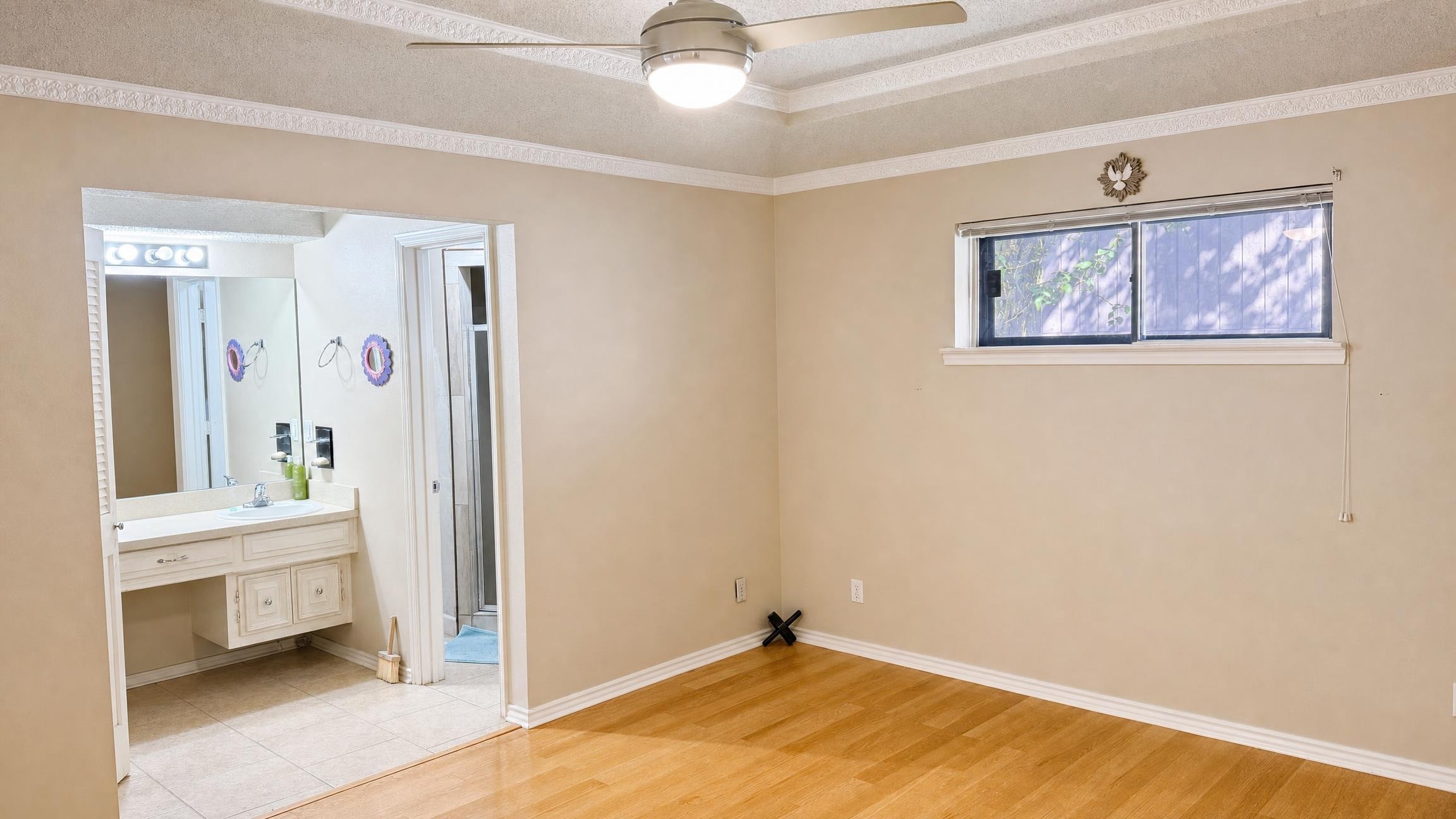 Empty minimal bedroom with beige walls, ceiling fan, hardwood flooring, and a small window above a baseboard with an attached bathroom vanity visible.