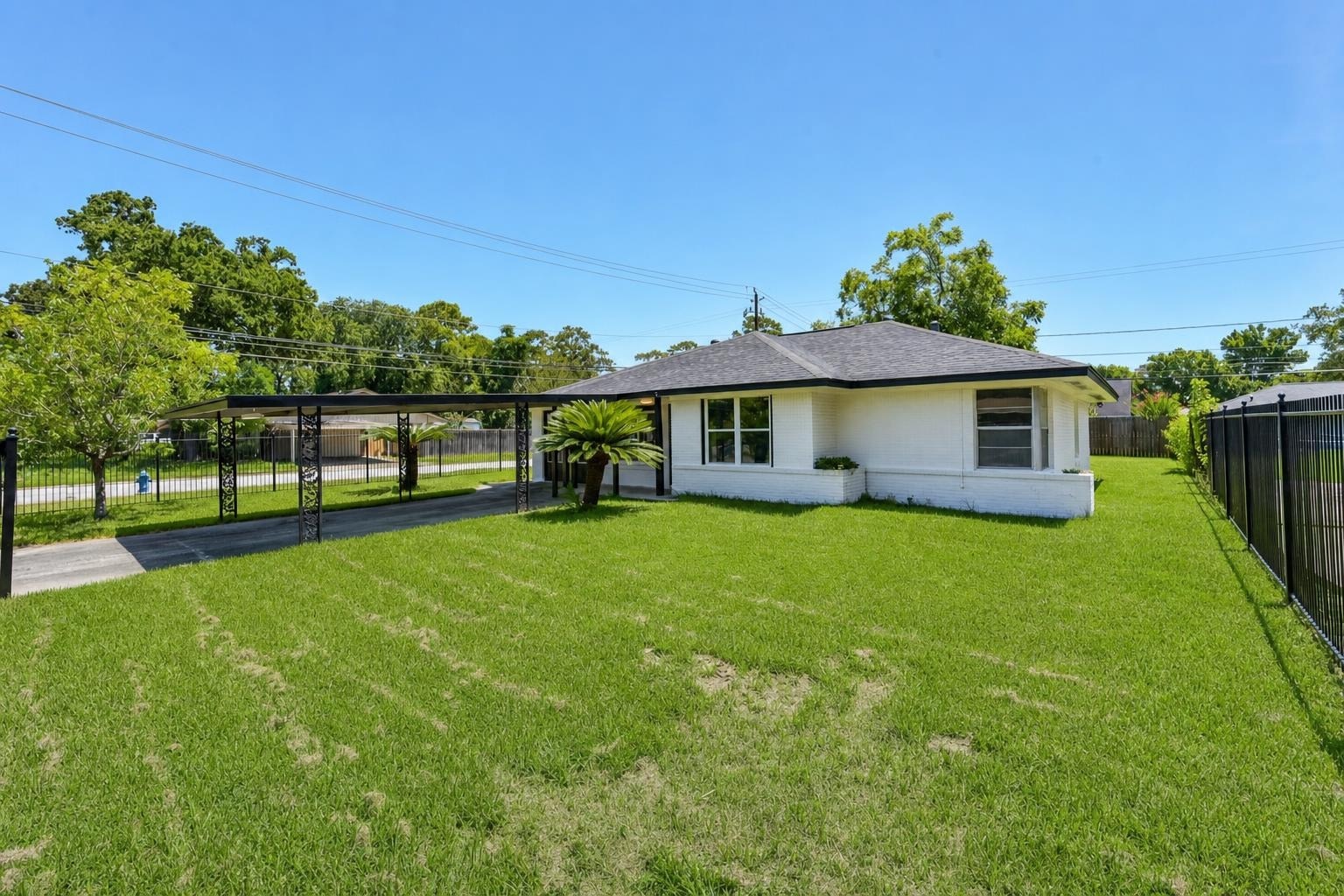 Single-story white house with dark roof, carport, palm tree, and large green lawn with fence and trees.