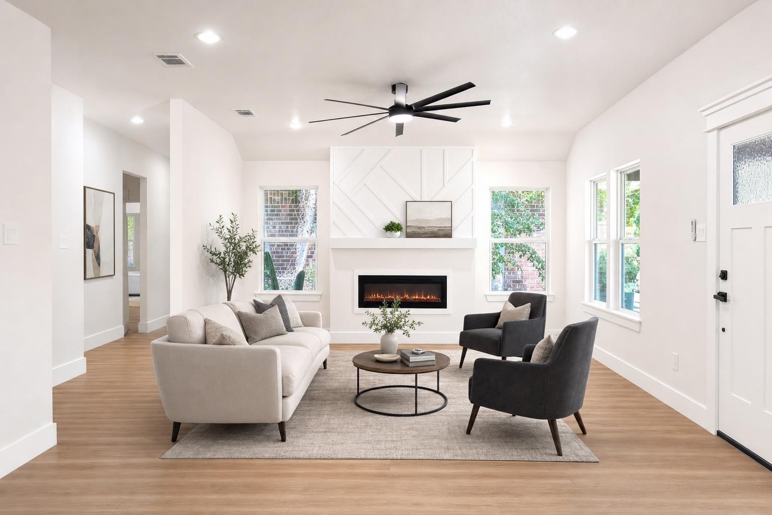 Modern living room with beige sofa, two charcoal armchairs, round coffee table, fireplace, and wood-look floor.