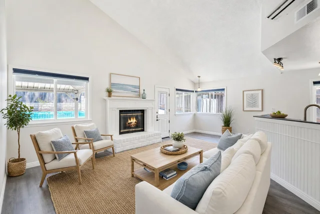 Bright coastal living room with white sofa, light wood chairs, coffee table, fireplace, and blue accent pillows.