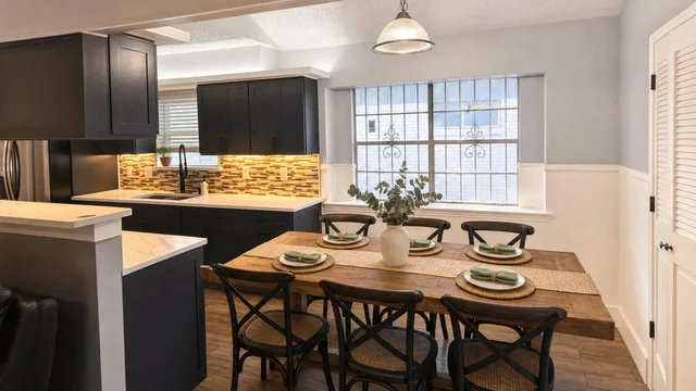 Dining room with large rustic wood table, six black cross-back chairs with woven seats, black kitchen cabinets, and a large window.