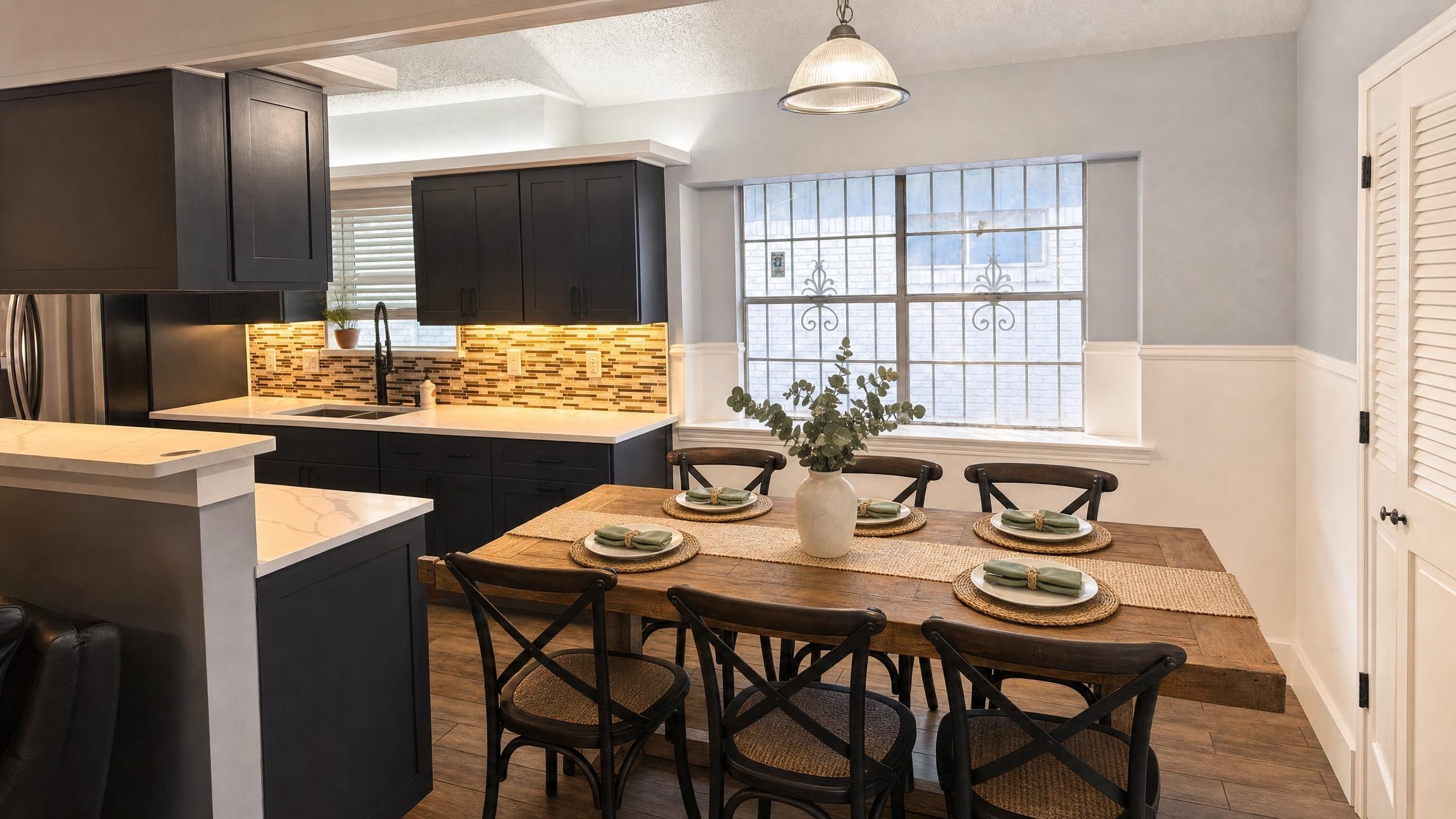 Dining room with large rustic wood table, six black cross-back chairs with woven seats, black kitchen cabinets, and a large window.