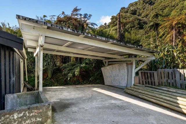 White wooden carport structure in front yard with concrete ground and green trees in background.