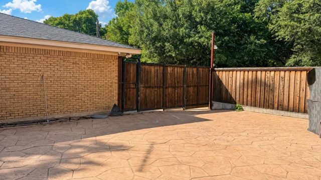 Backyard with stamped concrete floor, wooden fence gate, brick wall of a house, and green trees in the background.