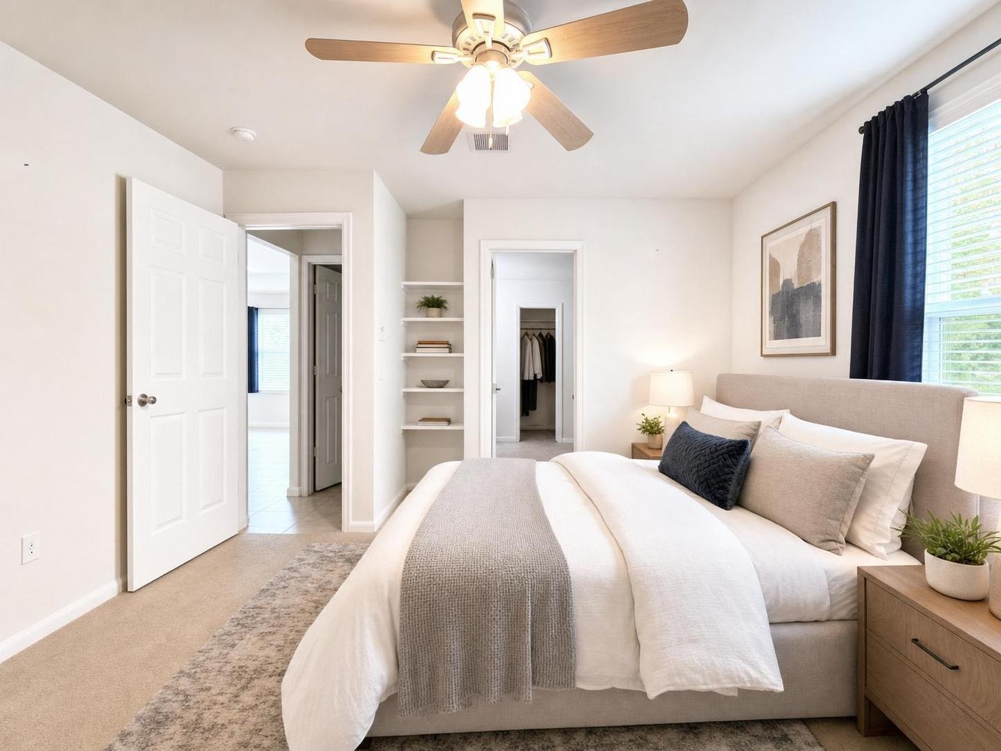 Bright bedroom with beige carpet, grey padded bed, wooden nightstands, white walls, and a ceiling fan with lights.