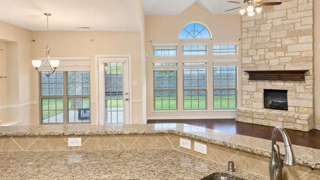 View from kitchen counter looking into living room with large windows, stone fireplace, and hardwood floor.