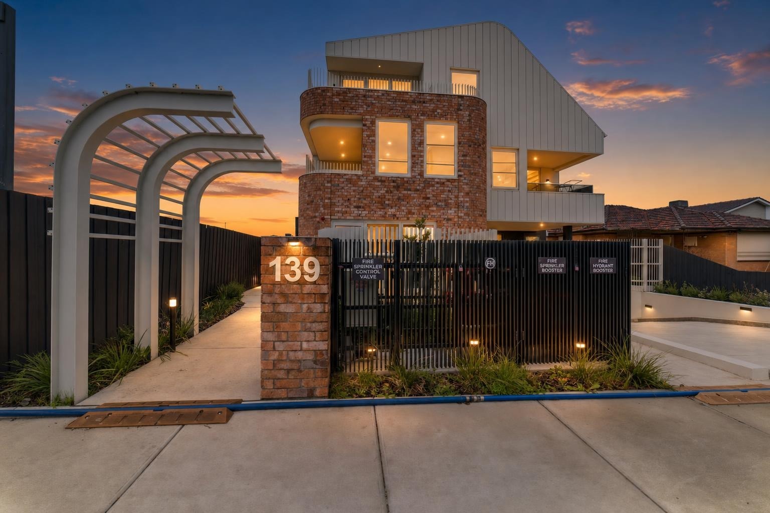 Modern three-story brick and metal house exterior with a black fence and illuminated house number 139 at dusk.