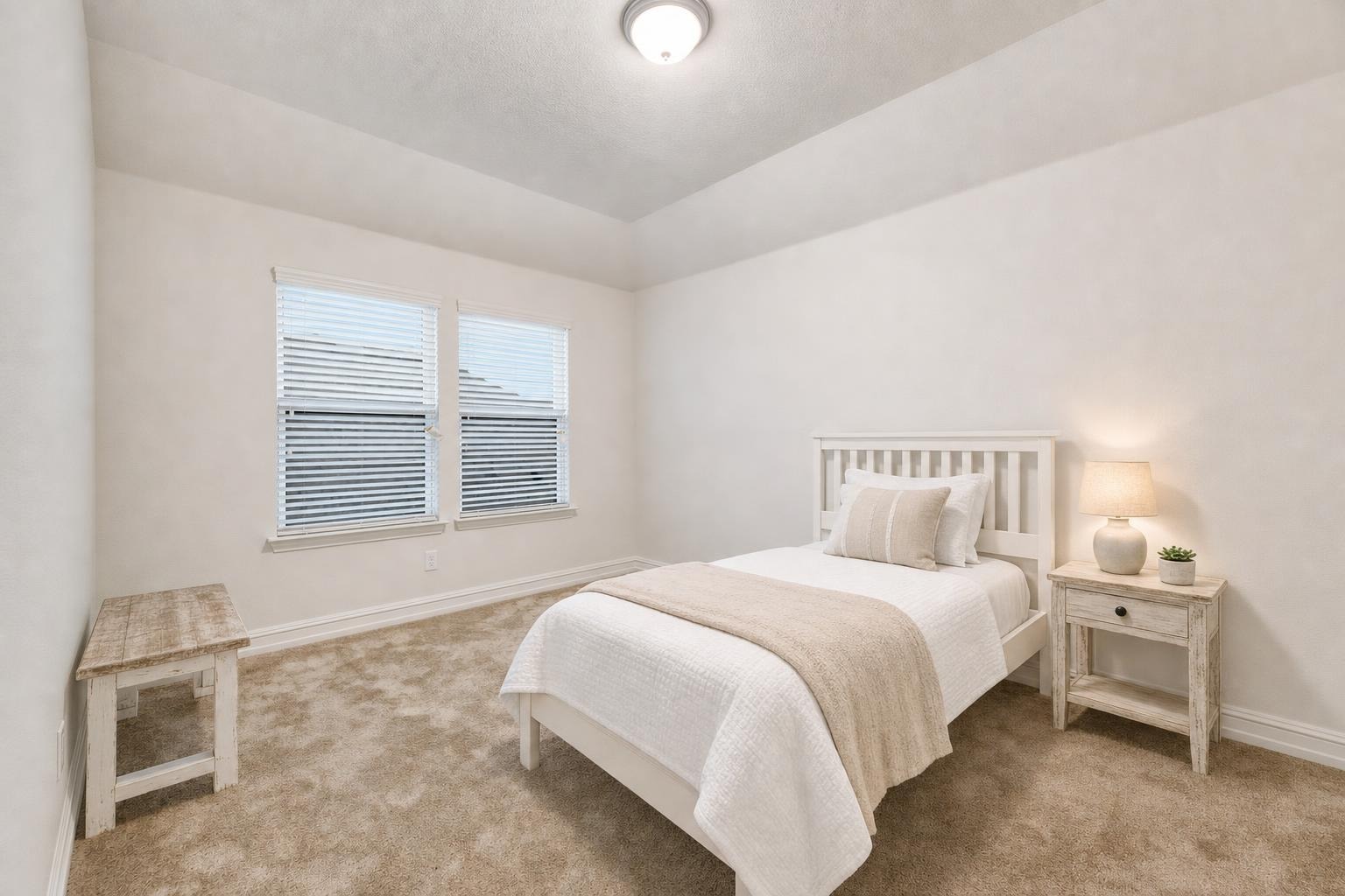 Farmhouse bedroom with white wooden bed, beige carpet, rustic wood bench, side table, lamp, and neutral bedding.
