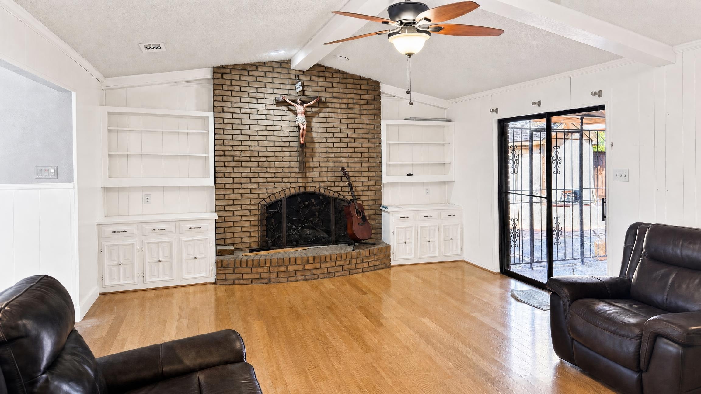 Living room with brick fireplace centered between white built-in shelves, two dark leather recliners, ceiling fan, and glass sliding door.