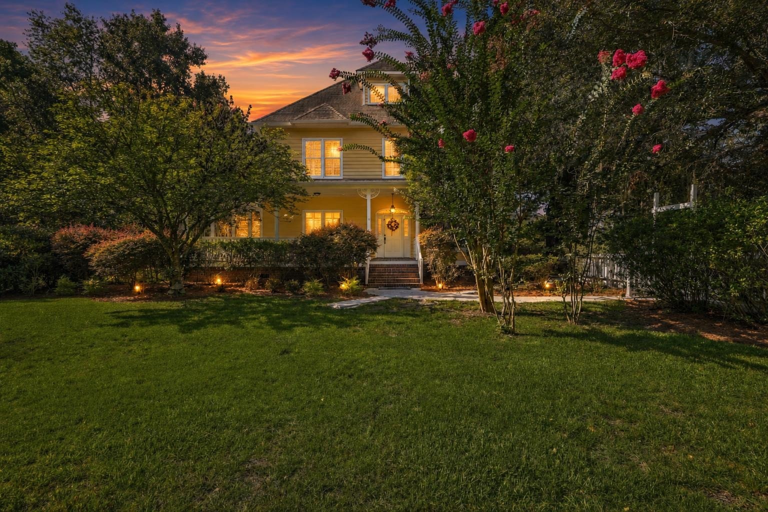 Traditional two-story yellow house with front porch, lit windows, trees, and green lawn at sunset.