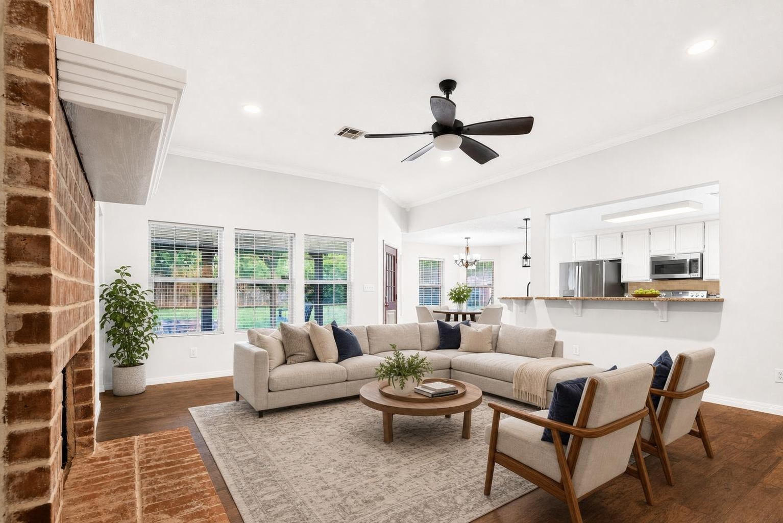Modern living room with beige sectional sofa, two beige armchairs with wooden frames, round wooden coffee table, large neutral area rug, and ceiling fan.