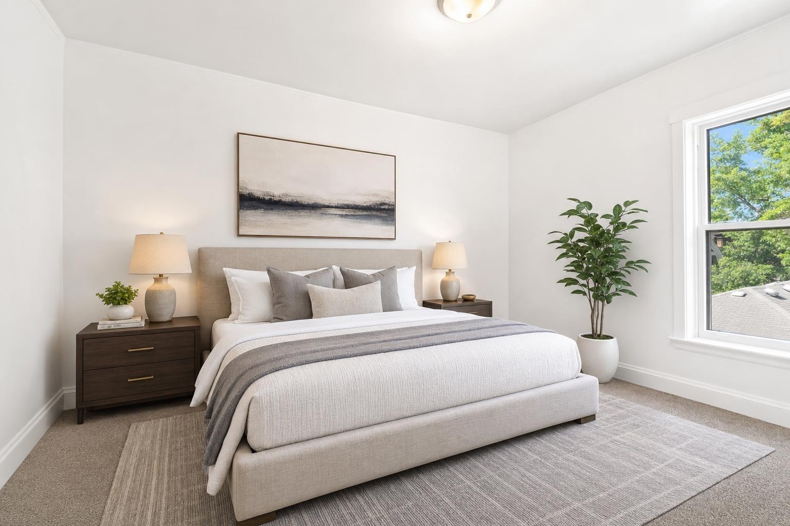 Bright bedroom with beige upholstered bed, two dark wood nightstands, table lamps, large window, and potted plant.