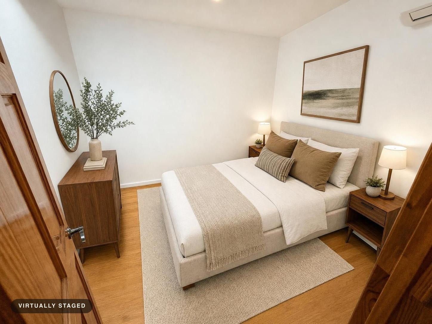 Modern bedroom with beige upholstered bed, neutral bedding, two wooden nightstands with lamps, wooden dresser, round mirror, and framed artwork.