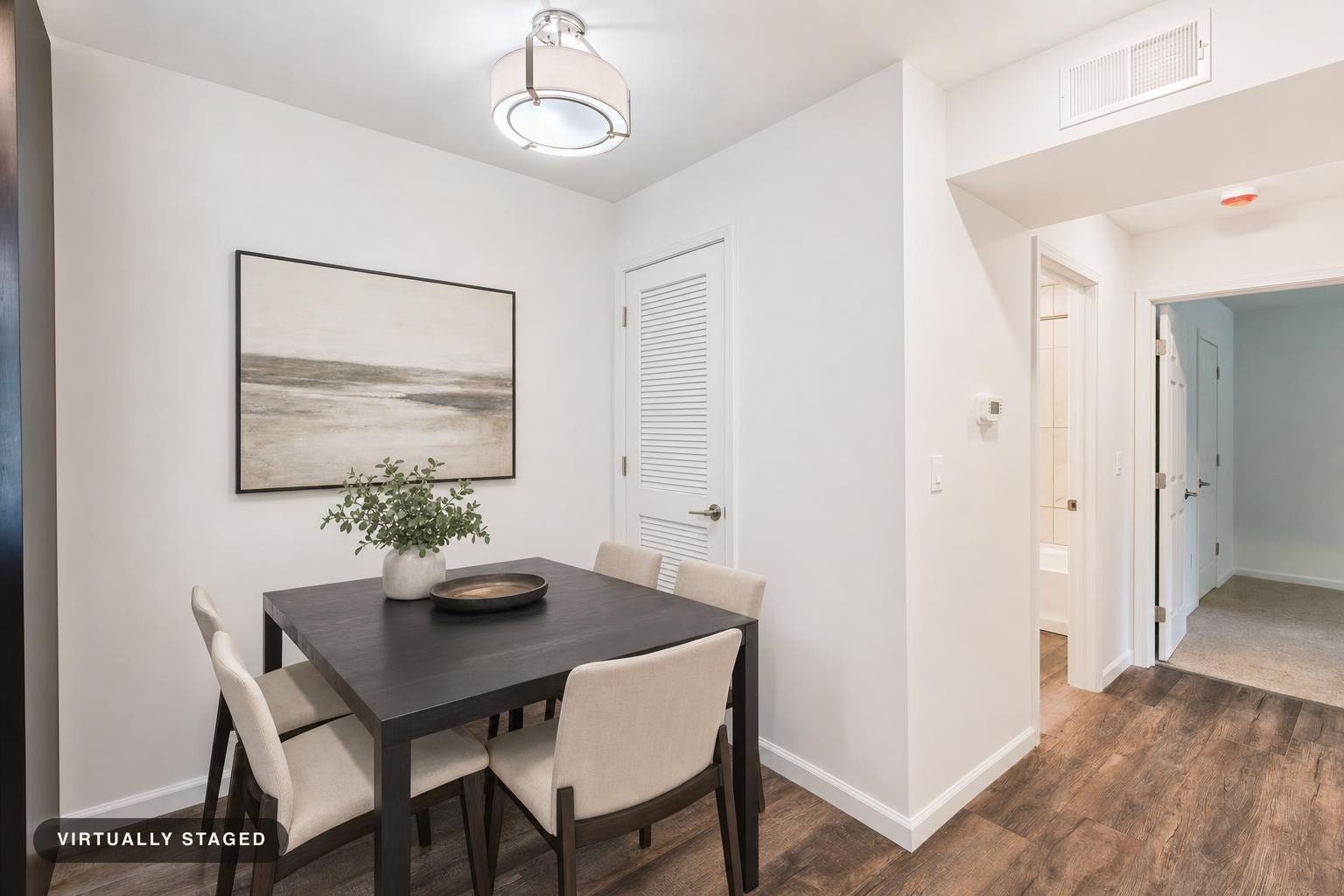 Dining room with black table, four beige chairs, wooden floor, white walls, and a framed landscape painting.