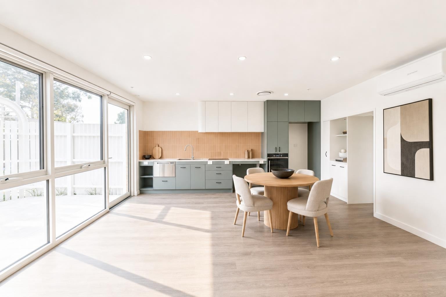 Modern dining room with round wooden table, four upholstered chairs, green and white kitchen cabinets, and large sliding glass doors.