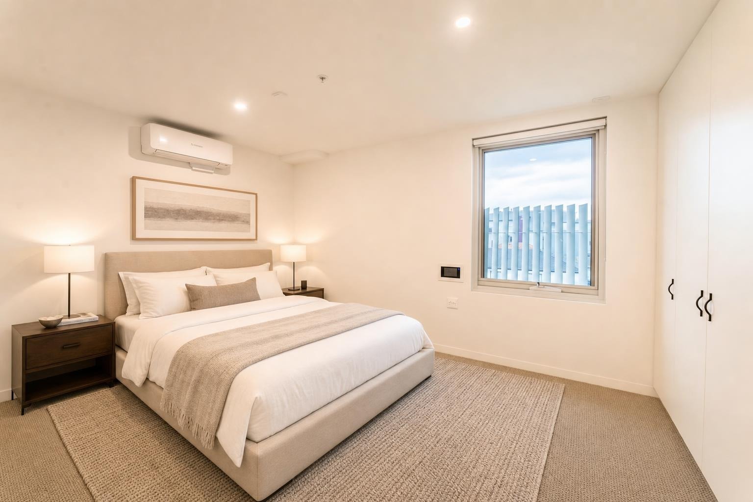 Modern bedroom with beige upholstered bed, two dark wood nightstands, wall art, window, and built-in white wardrobe.