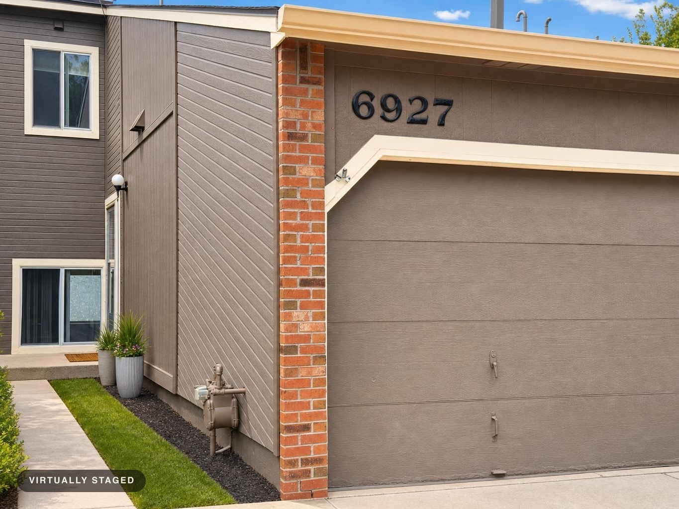 Front yard of modern home with taupe garage door, brick column, concrete pathway, and two large planters by the side door.