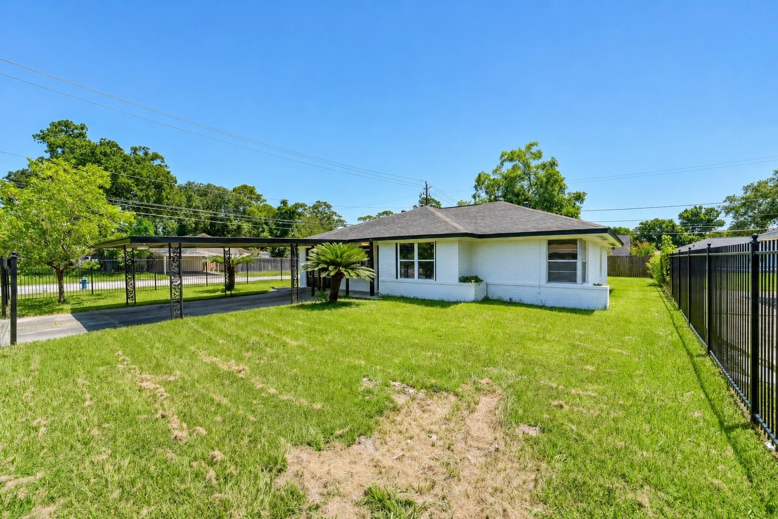 White single-story house with black roof, large green grassy yard, metal carport, and black fence on a sunny day.