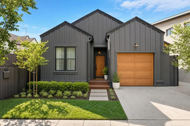Modern charcoal gray house exterior with vertical siding, wood front door and garage, small trees, and green lawn.