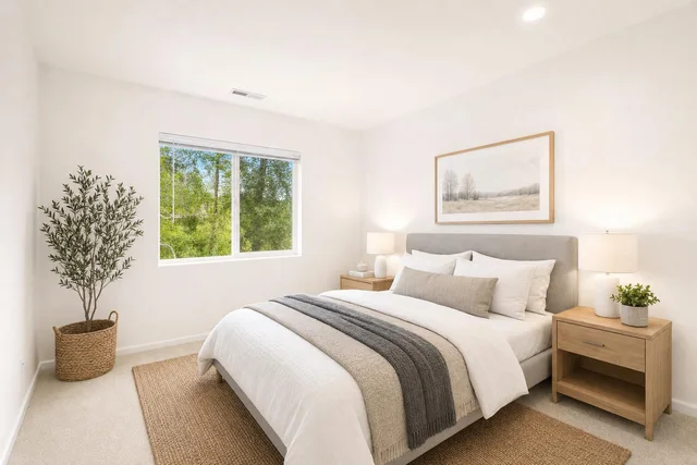 A bright bedroom with a queen bed dressed in white and beige bedding, flanked by light wood nightstands with lamps and a woven basket plant by the window.