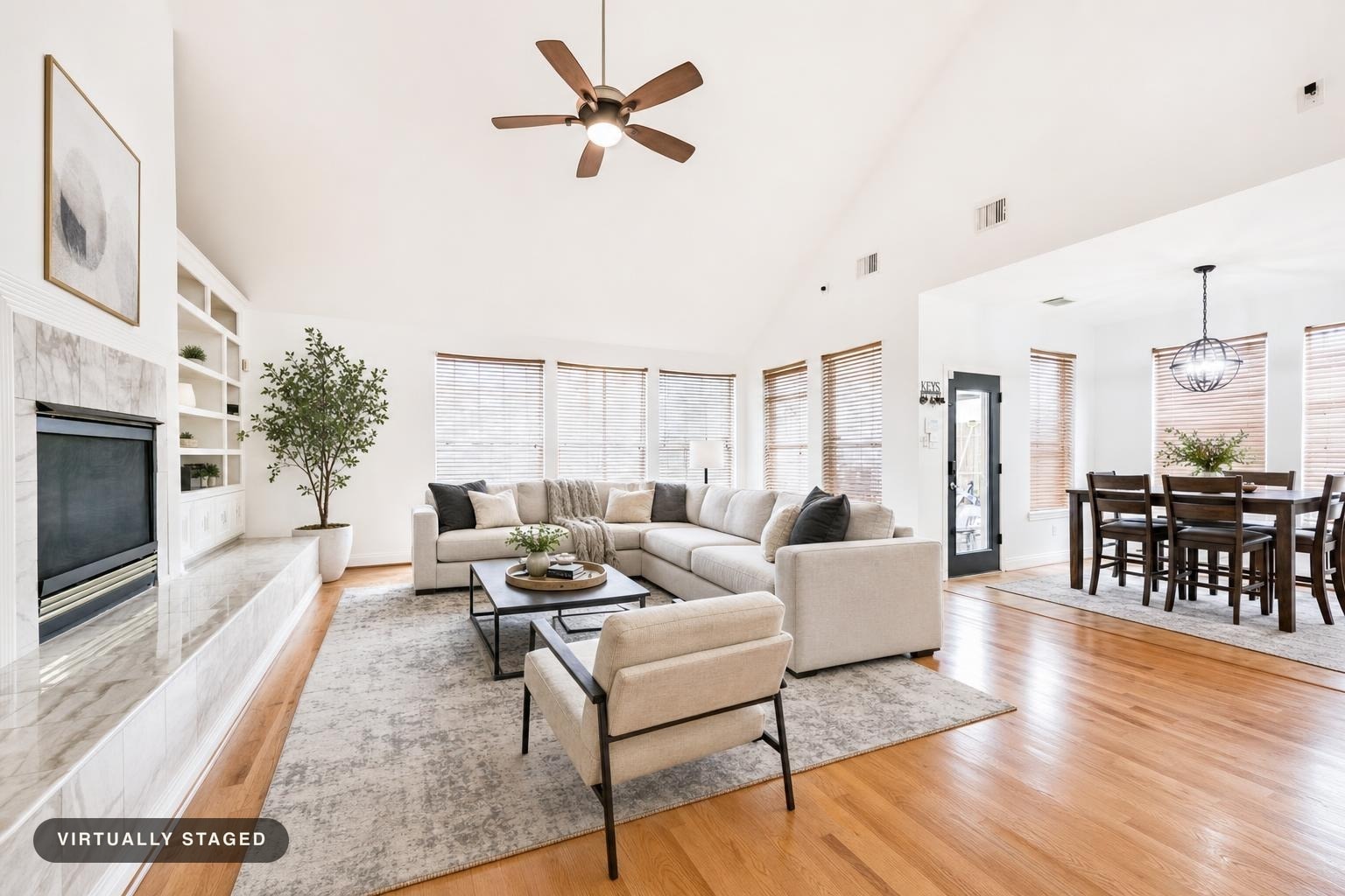 Bright living room with large beige sectional sofa, single armchair, black metal coffee table, wooden floor, and dining area visible.