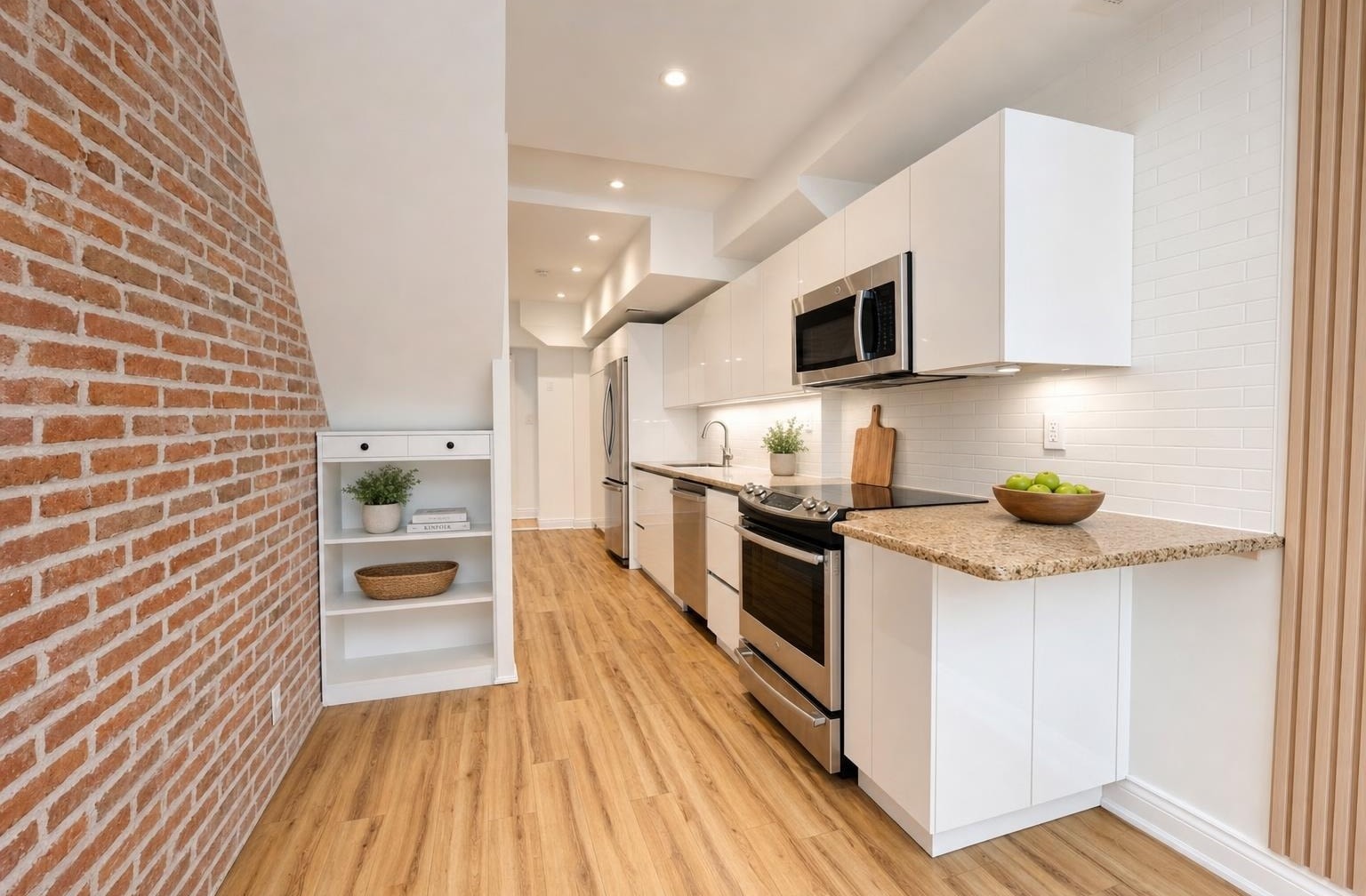 Modern kitchen with stainless steel oven and microwave, white cabinets, brick wall on left, and wood flooring.