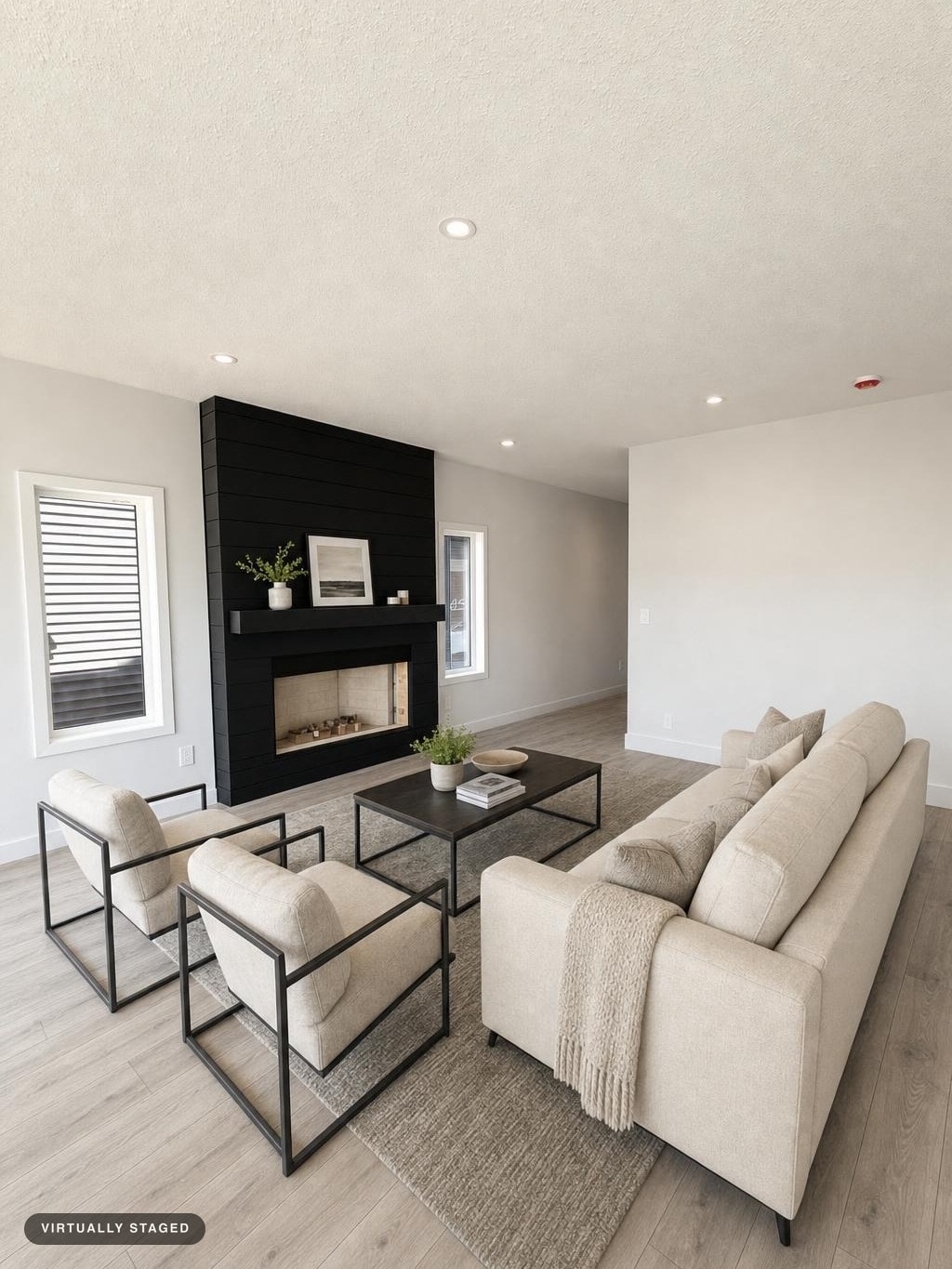 Modern living room with beige sofa, two black metal frame armchairs, rectangular coffee table, and black shiplap fireplace wall.