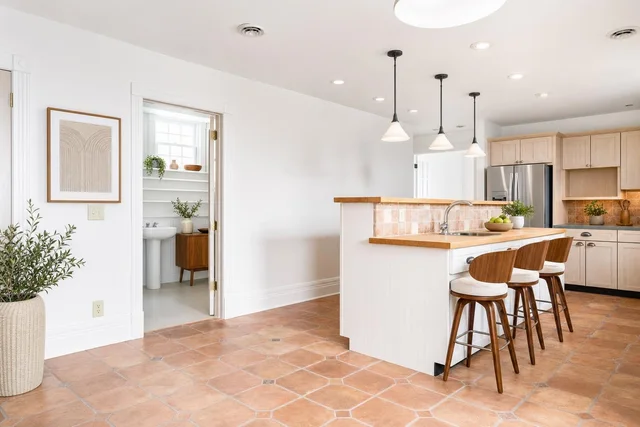 Bright modern kitchen featuring light wood cabinets, white island with wood countertop, three wood and white cushioned bar stools, and tiled floor.