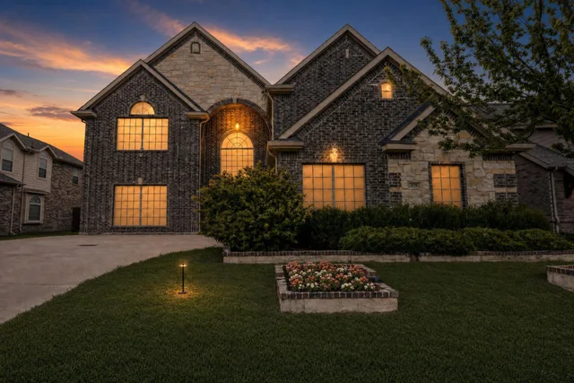 Two-story modern brick and stone house with warm lights on, landscaped front yard with flower bed and trimmed bushes at dusk.