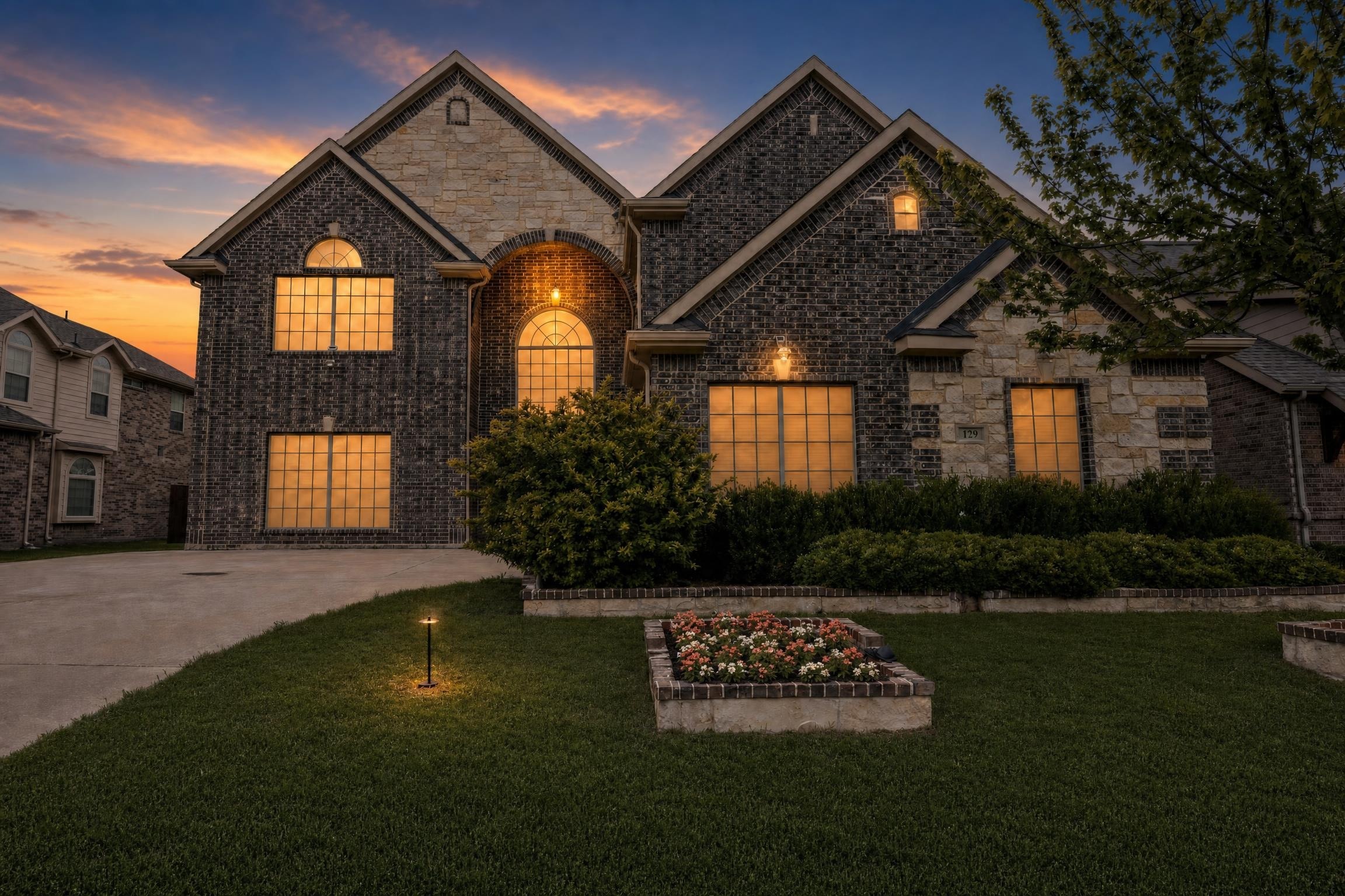 Two-story modern brick and stone house with warm lights on, landscaped front yard with flower bed and trimmed bushes at dusk.