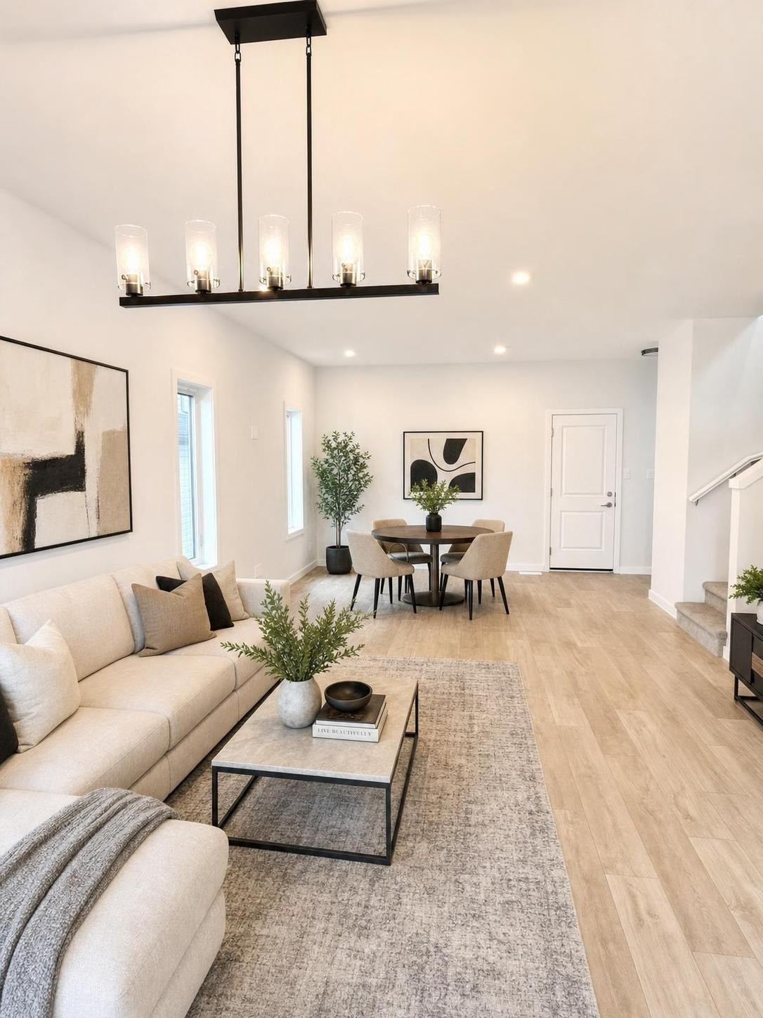 Modern living room with beige sofa, black-framed coffee table, abstract art, dining table with four chairs, and black linear chandelier.