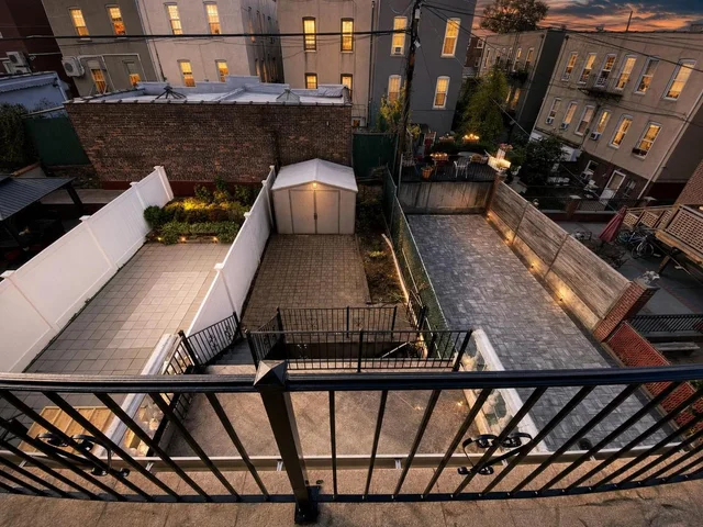 View of a modern backyard at dusk featuring paved areas, a small garden, a white shed, and black metal fencing.