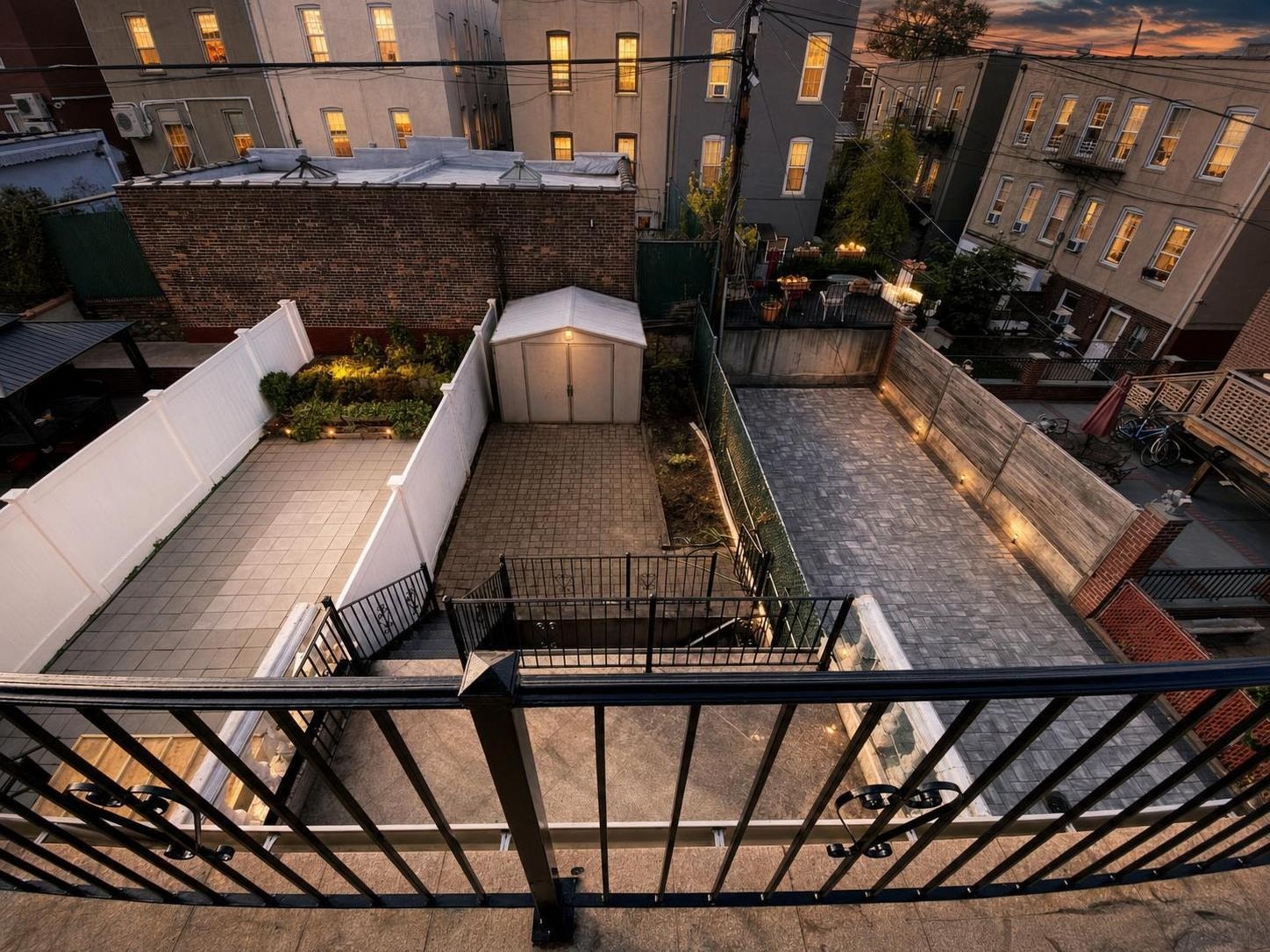 View of a modern backyard at dusk featuring paved areas, a small garden, a white shed, and black metal fencing.