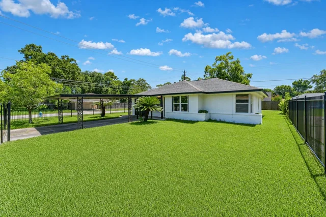 Small white house with gray roof and large green grass front yard, black metal carport, and blue sky with clouds.