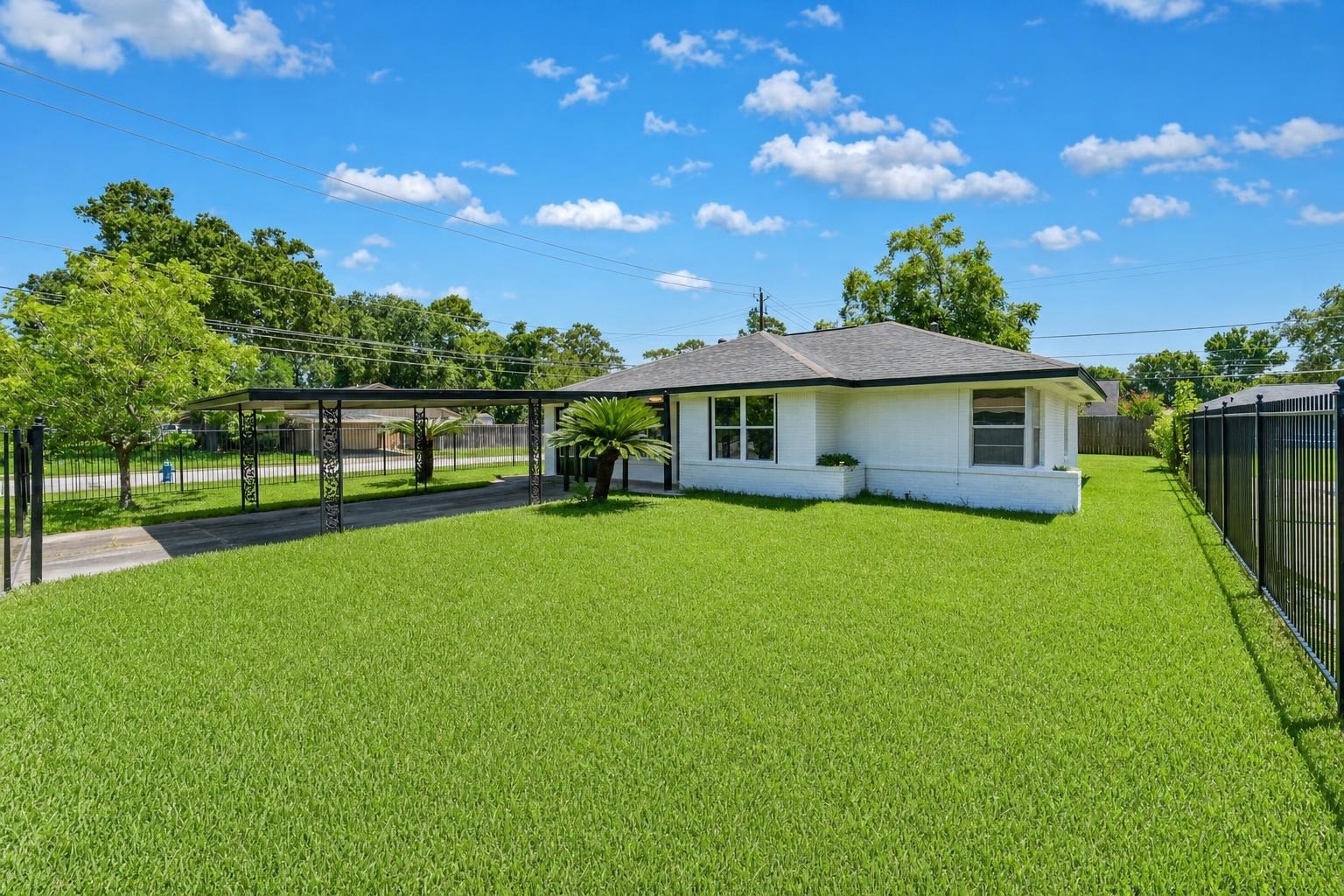 Small white house with gray roof and large green grass front yard, black metal carport, and blue sky with clouds.