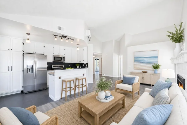 A bright coastal style living room with white walls, light wood furniture, blue cushions, and a kitchen with stainless appliances.