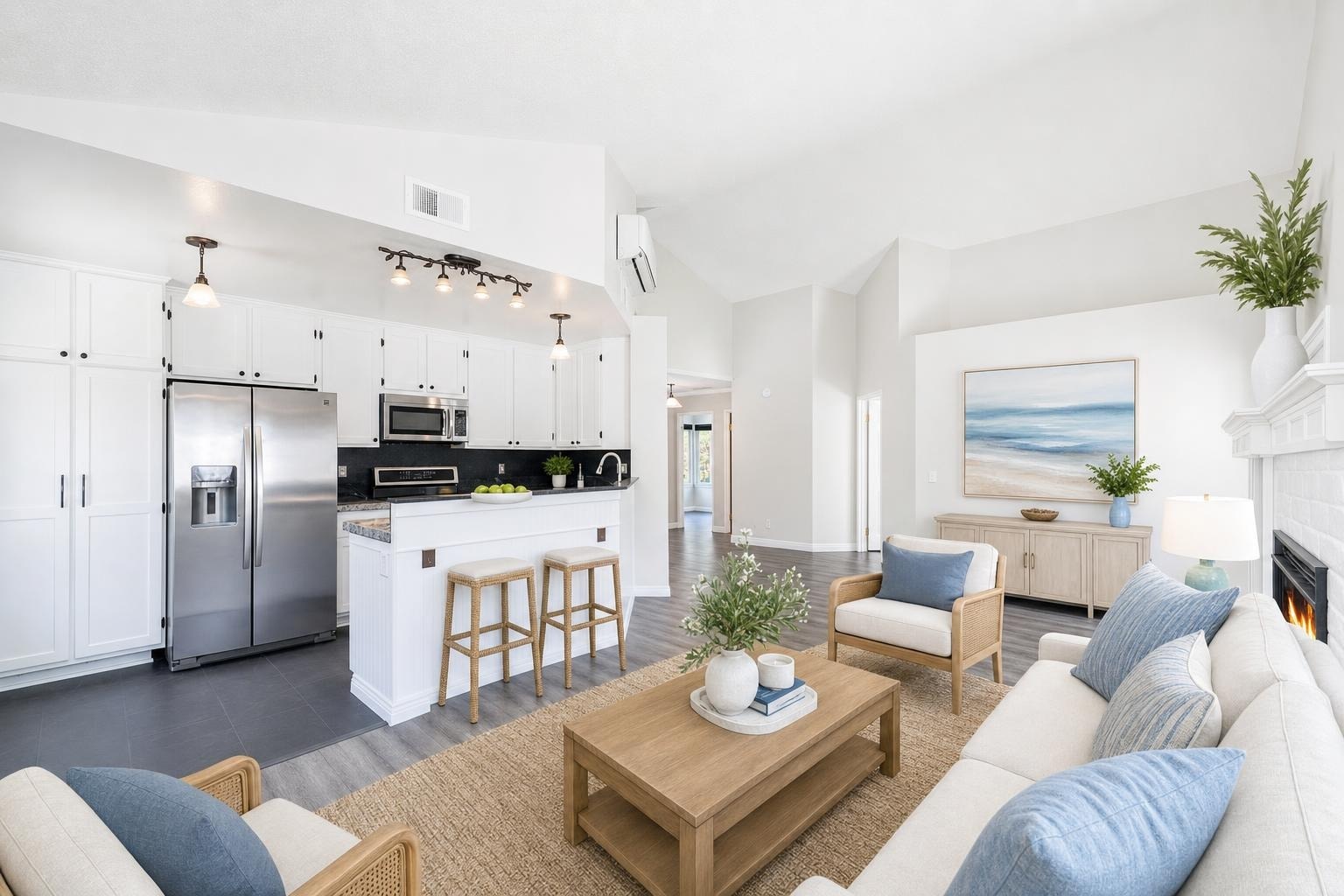 A bright coastal style living room with white walls, light wood furniture, blue cushions, and a kitchen with stainless appliances.